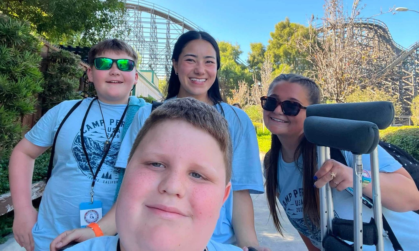 Four smiling Camp Perthes friends together on a sunny amusement-park field-trip day, roller coaster behind them, one using forearm crutches
