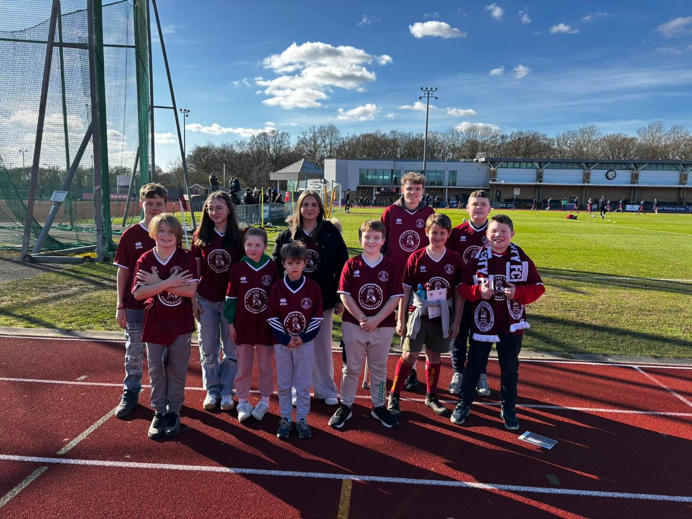 A group of Perthes Kids Foundation children in maroon PKF jerseys on a stadium athletics track on a bright sunny day out at a football club