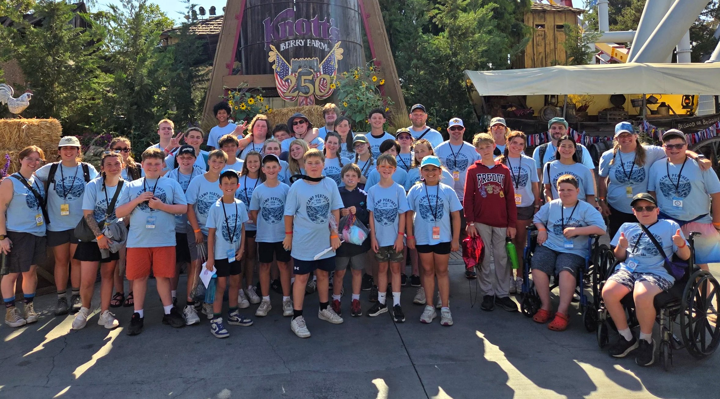 A large Camp Perthes group in light-blue camp shirts smiling together at Knott's Berry Farm, including many boys and two campers who use wheelchairs giving a thumbs up front right