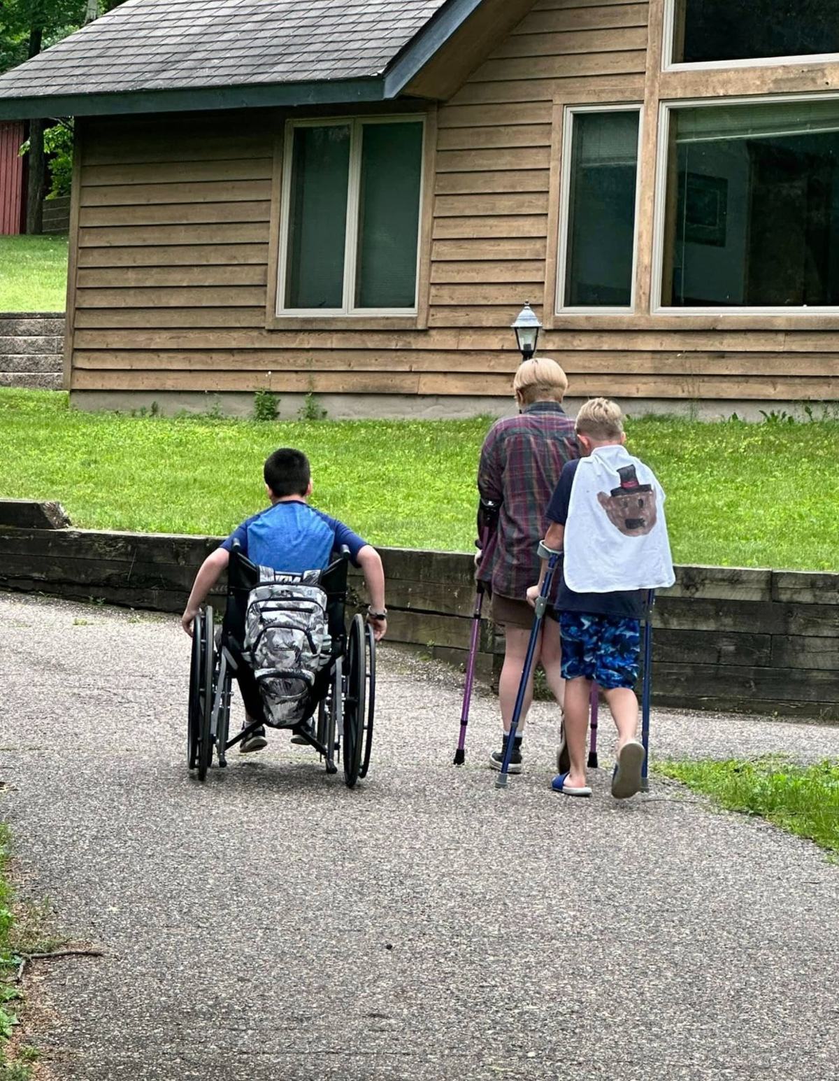 Three Camp Perthes kids seen from behind heading to a cabin — one using a wheelchair, one on forearm crutches, and one wearing a hand-drawn superhero cape