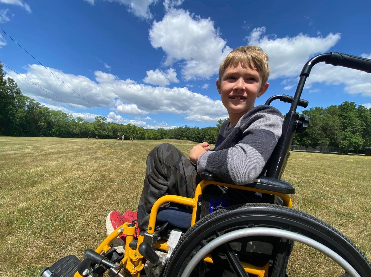 A smiling boy in a yellow wheelchair outdoors on a bright blue-sky day at camp